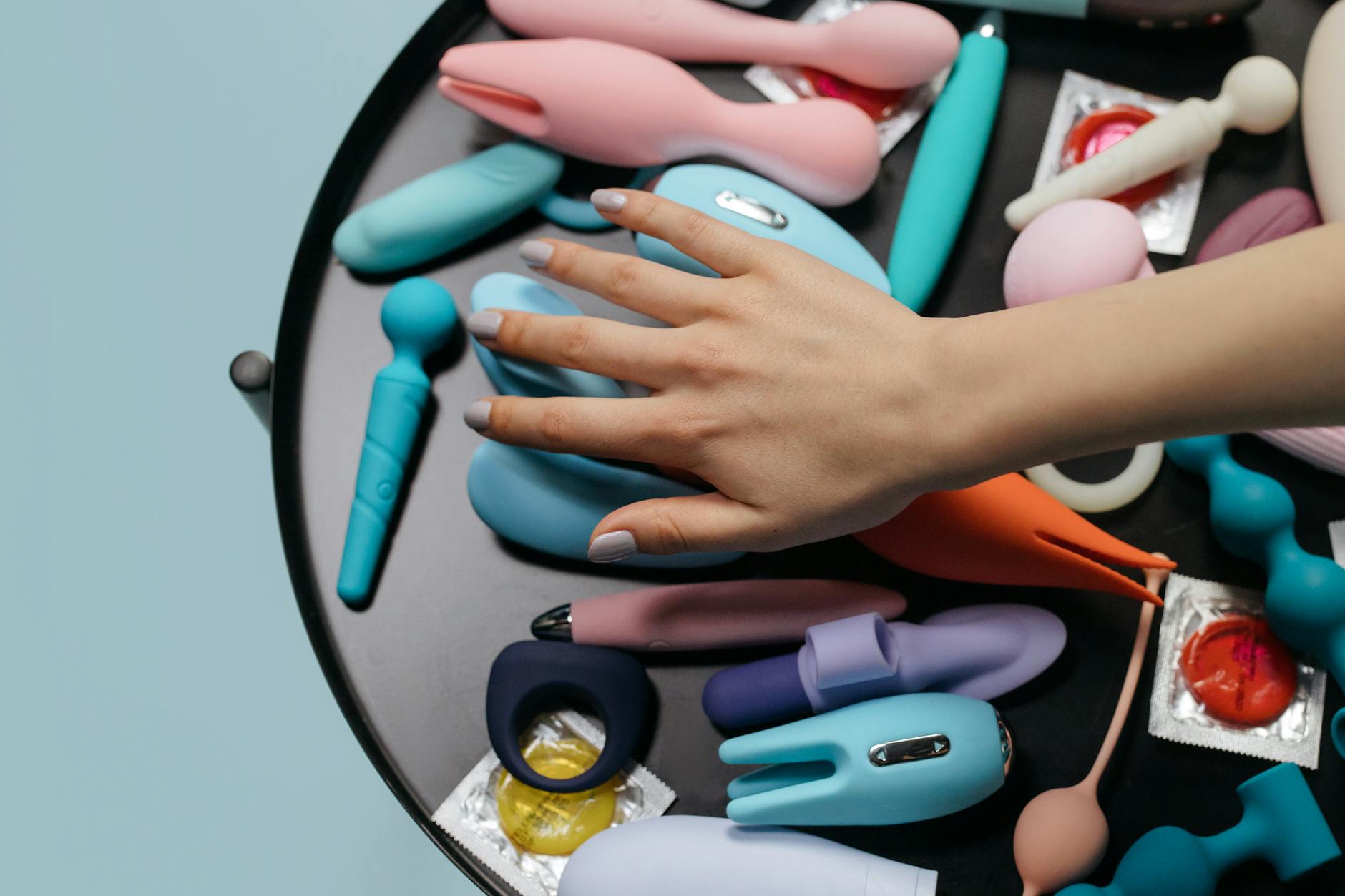 Hand reaching over a collection of colorful vibrators and personal massagers displayed together.